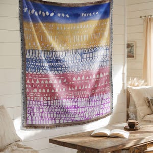 May include: A rectangular throw blanket with a colorful, abstract design. The blanket features horizontal bands of blue, yellow, red, and purple with geometric patterns. The edges are finished with a fringe.