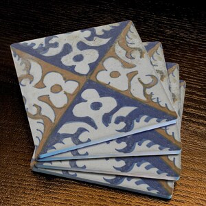 May include: A stack of square coasters with a floral design. Each coaster features a repeating pattern of white flowers, blue accents, and tan borders. The coasters are stacked on a dark wooden surface.