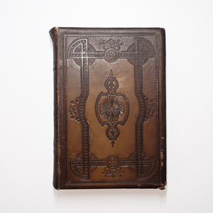 以下が含まれることがあります： 表紙に複雑な金色のエンボス加工が施された、ビンテージの茶色の革装丁の本です。