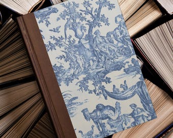 May include: A hardcover book with a blue and white toile pattern cover featuring a classical scene with figures, trees, and animals. The spine is a dark brown color. The book is surrounded by other open books.