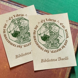 Puede incluir: Dos ex libris de papel marrón con ilustraciones verdes de un hombre y una mujer. El texto en los ex libris dice: "Ex Libris: Come, bebe y sé feliz, porque mañana moriremos." y "Ex Libris: Serp, porque mañana moriremos." La parte inferior de los ex libris dice: "Biblioteca" y "Biblioteca Daelli."