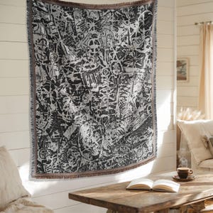 May include: A black and white woven throw blanket with a complex, abstract design. The pattern features various shapes and symbols, creating a textured appearance. The blanket has a fringed edge and is displayed on a white wall.