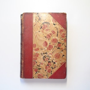 May include: A vintage book with a red leather spine and marbled paper cover. The cover features a pattern of red and black swirls on a cream background.