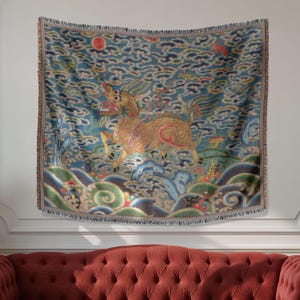 Op de afbeelding: Een decoratieve sprei met een op Azië geïnspireerd ontwerp. De sprei heeft een gouden mythisch wezen tegen een blauwe achtergrond met wolken- en golfpatronen. De randen hebben een franjes.