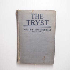 May include: A hardback book titled The Tryst by Grace Livingston Hill (Mrs. Lutz) with a light grey cloth cover. The title is printed in dark blue capital letters with two horizontal lines underneath, and the author name is below the title.