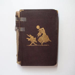 Puede incluir: Un libro antiguo con una cubierta marrón oscuro e ilustraciones doradas en relieve de dos figuras. El lomo del libro está desgastado y las páginas se sujetan con cinta adhesiva. La portada presenta una figura bailando y un músico.