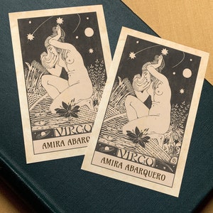 May include: Two vintage tarot cards with a black and white illustration of a woman with long hair, sitting with her hands on her head. The cards are on a dark green surface. The text on the cards reads "Virgo Amira Abarquero".