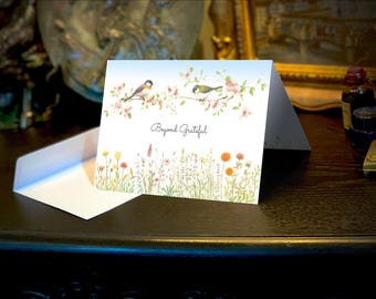 May include: A white card with a blue sky background and a floral design. Two birds are perched on branches with pink flowers. The text "Beyond Grateful" is printed in black on the card.