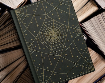 Könnte beinhalten: Ein dunkelgrünes Buch mit einem goldenen Spinnennetz-Design und Sternakzenten. Das Hardcover-Buch liegt auf einem Hintergrund aus offenen Büchern. Das Spinnennetz-Design ist mittig auf dem Einband platziert.