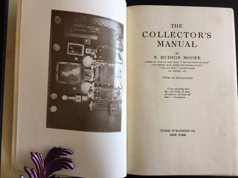May include: An open book titled "The Collector's Manual" by N. Hudson Moore. The book features a sepia-toned illustration and text. Published by Tudor Publishing Co. in New York. The book's pages are off-white.