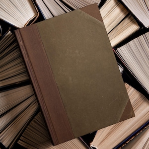 Puede incluir: Un libro de tapa dura con una cubierta y lomo marrones, sobre un fondo de libros abiertos. La cubierta del libro es de un verde oliva apagado, con un lomo y esquinas de color marrón oscuro. Las páginas de los libros circundantes están envejecidas y amarillentas.