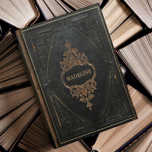 May include: A dark green book with gold detailing and the name "MADELINE" in a gold frame. The book is surrounded by other books with open pages, creating a vintage aesthetic. The cover has an embossed design.