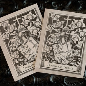 May include: Two black and white prints of a coat of arms with the name "Dominique Carrell" at the bottom. The coat of arms features a shield with a cross and a crown, surrounded by floral designs.