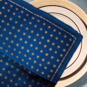 May include: A navy blue napkin with a repeating pattern of gold stars. The napkin is placed on a white plate with a brown rim, which sits on a wooden charger. The napkin has a decorative border.