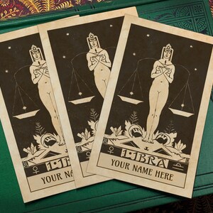 May include: Three vintage-style tarot cards with a black and white illustration of a woman holding a scale. The cards are on a green and brown surface. The text on the cards reads "Libra" and "Your Name Here".