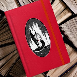 May include: Red notebook with a black and white illustration of a woman and a figure with wings, surrounded by a snake, inside a black oval. The notebook has a red spine.