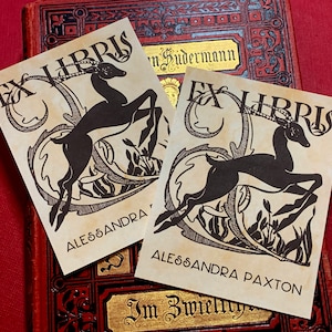 Puede incluir: Dos ex libris antiguos con una ilustración en blanco y negro de un antílope con un borde decorativo. El texto "Ex Libris" está impreso en negro en la parte superior de cada ex libris. El nombre "Alessandra" está impreso en la parte inferior del primer ex libris y "Alessandra Paxton" está impreso en la parte inferior del segundo ex libris.
