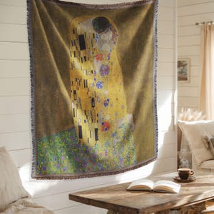 Op de afbeelding: Een geweven sprei met Gustav Klimt's kunstwerk "De Kus". De sprei toont het iconische beeld van het omarmende paar in goud-, geel- en zwarttinten, met een franjerand. Het kunstwerk is geplaatst tegen een bruine achtergrond.