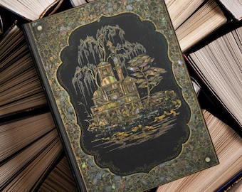May include: A decorative black book with an ornate, iridescent design featuring a building and trees. The book is surrounded by open books, suggesting a literary theme. The cover has a detailed, inlaid design with gold and iridescent accents.