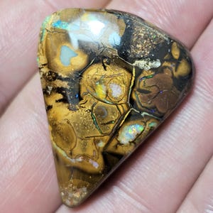 Può includere: Una gemma di opale lucidata, di forma triangolare. La pietra presenta un mosaico di colori, tra cui oro, marrone, nero e lampi iridescenti di blu, verde e giallo. La superficie dell'opale ha un aspetto unico e strutturato, che mostra motivi naturali.