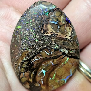 May include: A polished, teardrop-shaped opal gemstone. The stone features a mix of brown, black, and tan colors with iridescent flashes of green, blue, and purple. The opal has a unique, textured appearance.