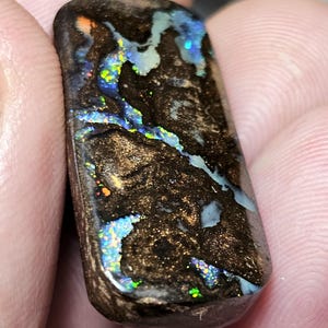 Könnte beinhalten: Ein rechteckiger, polierter Opal-Edelstein mit dunkelbrauner Grundfarbe. Der Opal weist lebhafte Blitze in Blau, Grün und Gelb auf, wodurch ein farbenfroher, schillernder Effekt entsteht. Der Stein wird zwischen den Fingern gehalten und zeigt seine natürliche Schönheit.