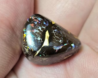 Puede incluir: Una piedra preciosa de ópalo pulido, de color marrón oscuro, con destellos iridiscentes. El ópalo tiene una superficie agrietada y texturizada con inclusiones doradas. La piedra preciosa se sostiene en una mano, mostrando sus patrones y colores únicos.