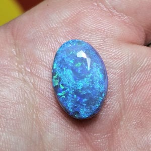 May include: An oval-shaped opal gemstone with a vibrant display of blue and green colors. The gemstone is held in a hand, showcasing its polished surface and internal play of color. The opal's unique pattern and iridescence are highlighted.
