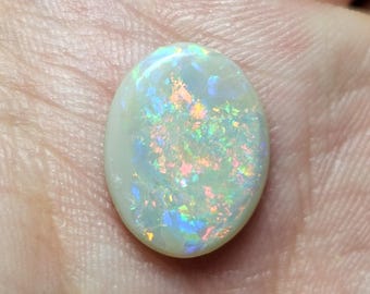 May include: Oval-shaped opal gemstone with a milky white base and vibrant flashes of color. The opal displays a play-of-color effect, with iridescent hues of blue, green, orange, and yellow. The gemstone is held in a hand.