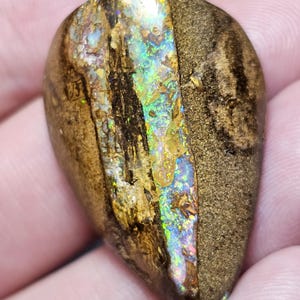 Può includere: Una gemma a forma di goccia con un esterno marrone e strutturato e un centro iridescente vibrante. Il centro mostra uno spettro di colori, tra cui verde, blu e oro, creando un contrasto visivo sorprendente. La pietra è tenuta in una mano.