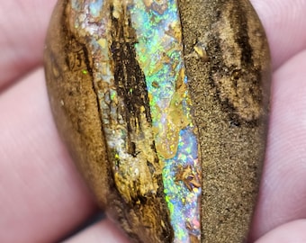 Puede incluir: Una piedra preciosa en forma de lágrima con un exterior marrón texturizado y un centro iridiscente vibrante. El centro muestra un espectro de colores, incluyendo verde, azul y dorado, creando un contraste visual llamativo. La piedra se sostiene en una mano.