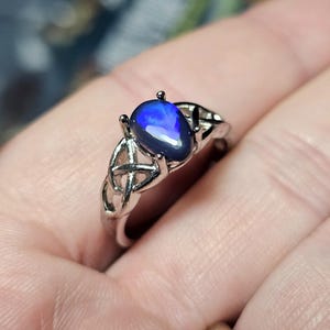 May include: A silver-toned ring featuring a pear-shaped, dark blue iridescent stone set in a four-prong mount. The band is designed with an intricate, openwork Celtic knot pattern that extends from the setting down the sides of the ring.