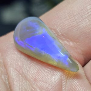 May include: A teardrop-shaped opal gemstone featuring a translucent, pale yellow base color with a vibrant, iridescent blue flash across the center. The stone has a smooth, polished surface and a curved, cabochon-style top profile.