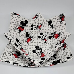 May include: White fabric bowl cozy with a repeating pattern of Mickey Mouse in black, red, and white. The cozy is square with folded corners, designed to hold a bowl. The background is white with small gray Mickey Mouse silhouettes.