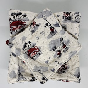 May include: Cream-colored, quilted bowl cozies featuring a repeating pattern of Mickey and Minnie Mouse in various scenes. The design includes red accents, gray clouds, and iconic landmarks. The cozies are square-shaped with a quilted texture.