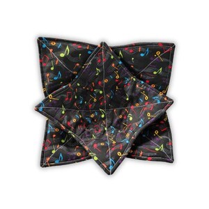 May include: A square fabric bowl cozy featuring a black background with a colorful pattern of musical notes and staff lines. The quilted item has four pointed corners and is constructed with visible stitching that creates a geometric grid pattern.