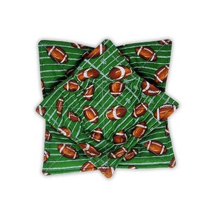 May include: A square fabric bowl cozy featuring a green field pattern with white yard lines and brown football graphics. The quilted textile accessory has a folded corner design and is constructed with visible stitching throughout the fabric.