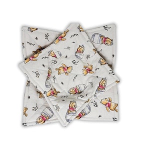 May include: Two square quilted fabric bowl cozies featuring an off-white background with a repeating watercolor-style print of a yellow bear in a red shirt, depicted in various poses including sitting in a blue pot, with small black sprig accents.