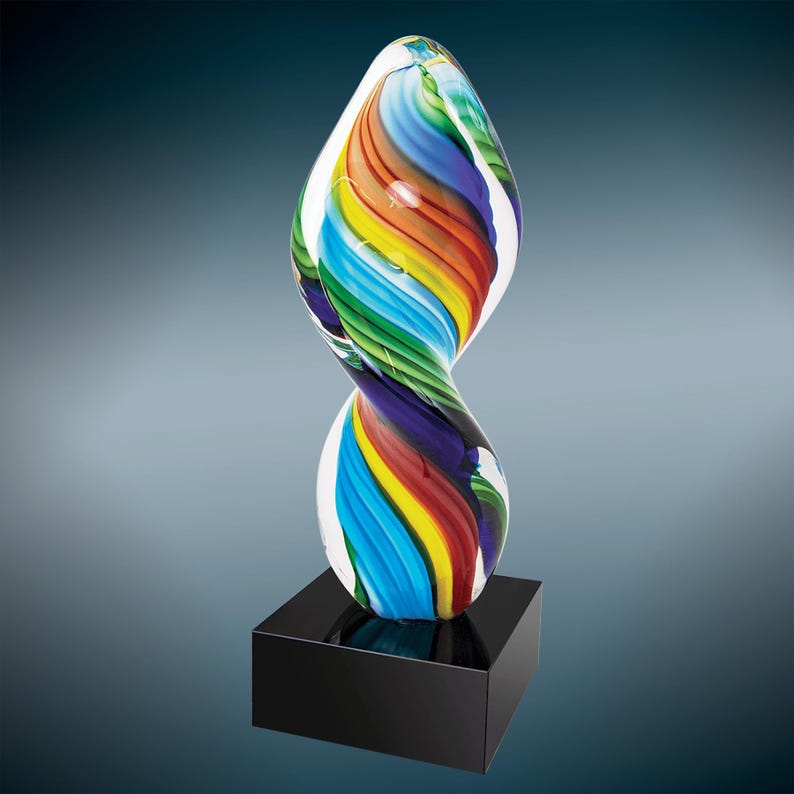 May include: A colorful glass sculpture with a twisted, flowing design. The sculpture features vibrant bands of red, orange, yellow, green, blue, and purple. It is mounted on a black square base, creating a striking contrast.