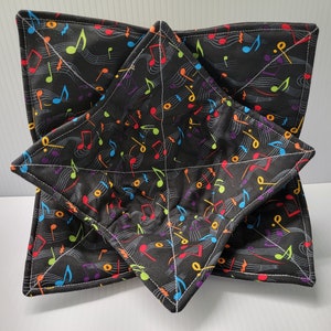 May include: Two square fabric bowl cozies, one nested inside the other. The black fabric is covered in colorful musical notes and staff lines. The edges are finished with white stitching. These cozies are designed to be used in the microwave.