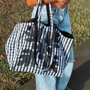 May include: A person wearing black sunglasses, a black crop top, and light blue jeans, holding a large black and white tote bag. The tote bag has a checkered pattern with a floral design. The person is also wearing hoop earrings and sandals.