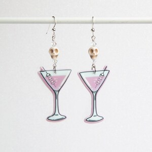 Peut inclure: Une paire de boucles d'oreilles argentées avec un charme en verre à martini rose et une perle de crâne blanche. Le verre à martini contient une boisson rose et une paille avec trois crânes blancs.