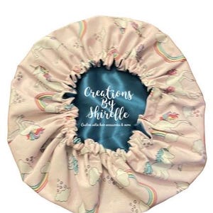 May include: A pink satin bonnet with a pattern of unicorns, rainbows, and clouds. The interior is teal satin. The text "Creations By Shirelle" is visible on the interior. Custom satin hair accessories & more.