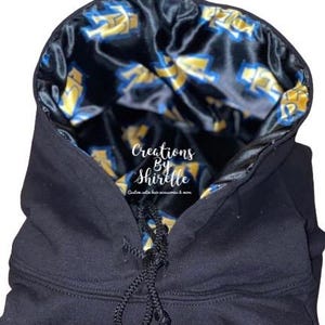 May include: Black hoodie with a satin lining featuring a pattern in yellow and blue. The hood has a drawstring closure. The text "Creations By Shirelle" is visible on the front.