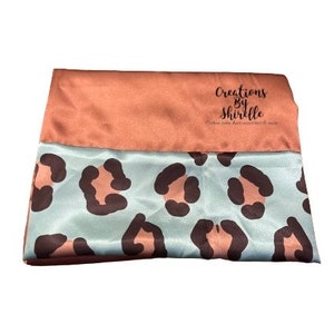 May include: A peach-colored satin fabric with a leopard print satin fabric sewn to the bottom. The fabric has the text "Creations By Shirtelle" printed on it.