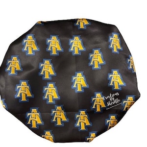 May include: Black satin bonnet featuring a repeating pattern of gold and blue collegiate logos. The bonnet is designed to protect hair and maintain hairstyles. The logo is a stylized "AT" with a blue outline.