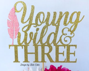 May include: Gold glitter cake topper with the words "Young Wild & Three" and a pink feather. The topper is designed for a third birthday party.