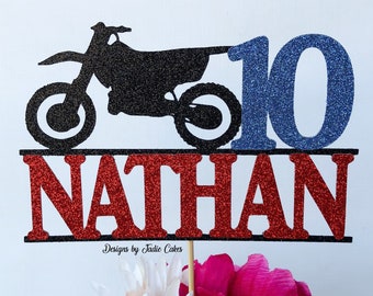 May include: A glittery black motorcycle cake topper with the number 10 in blue glitter and the name "NATHAN" in red glitter. The cake topper is on a wooden stick and is placed in front of a bouquet of flowers.