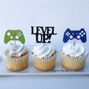 May include: Three cupcakes decorated with green, blue, and black glitter game controller toppers. The cupcake in the middle has a black glitter topper that says "LEVEL UP!"
