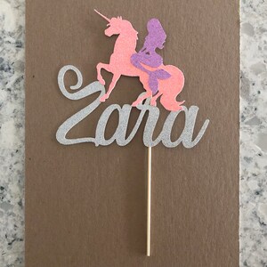 May include: A silver glitter cake topper with the name "Zara" written in cursive. A pink unicorn with a purple mermaid riding on its back.